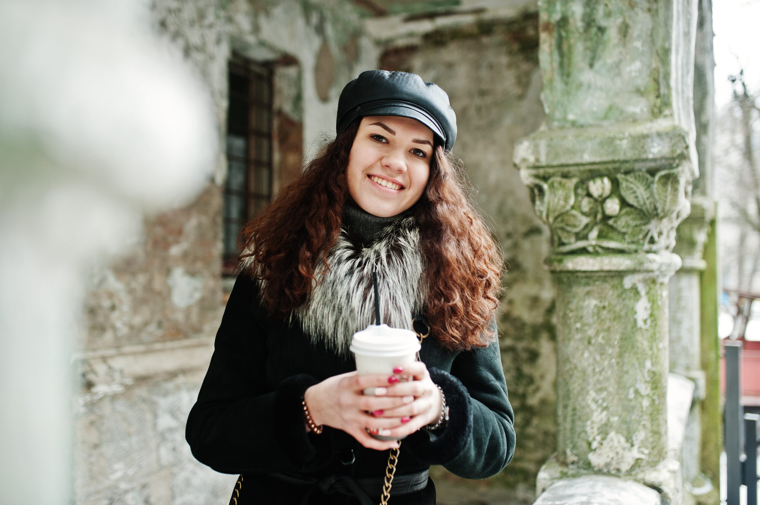 Curly mexican girl in leather cap and plastic cup of coffee at hand walking at streets of city.